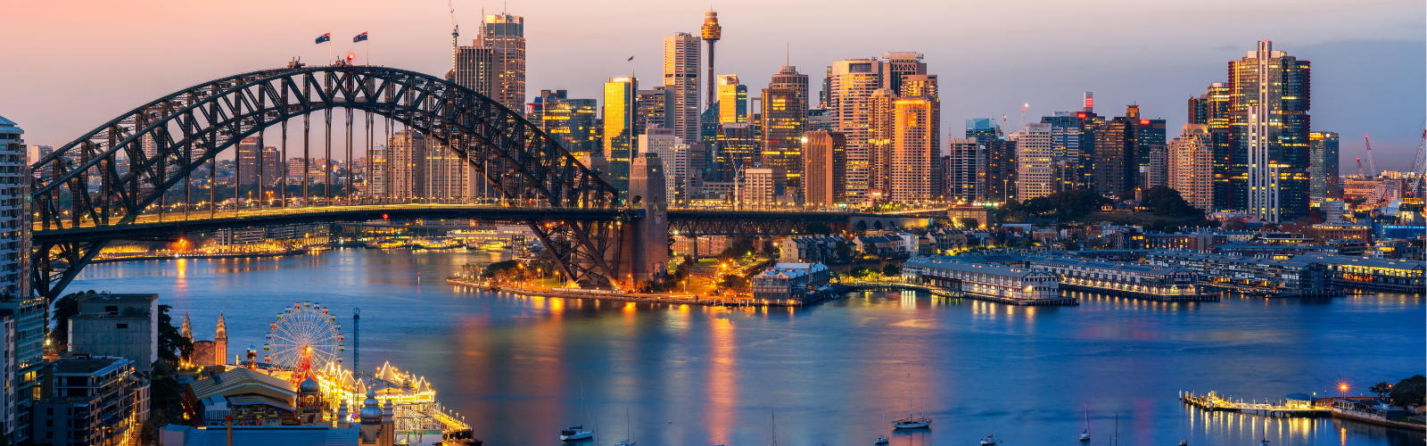 Sydney city skyline at dusk with Sydney Harbour Bridge spanning the harbor, high-rise buildings in the background, and water reflecting city lights in the foreground.